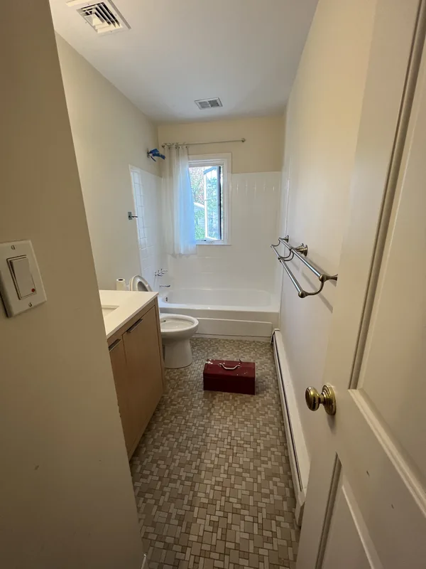 The original Great Neck bathroom before remodeling, seen from the doorway, with beige walls, vinyl flooring and a shower curtain over the tub