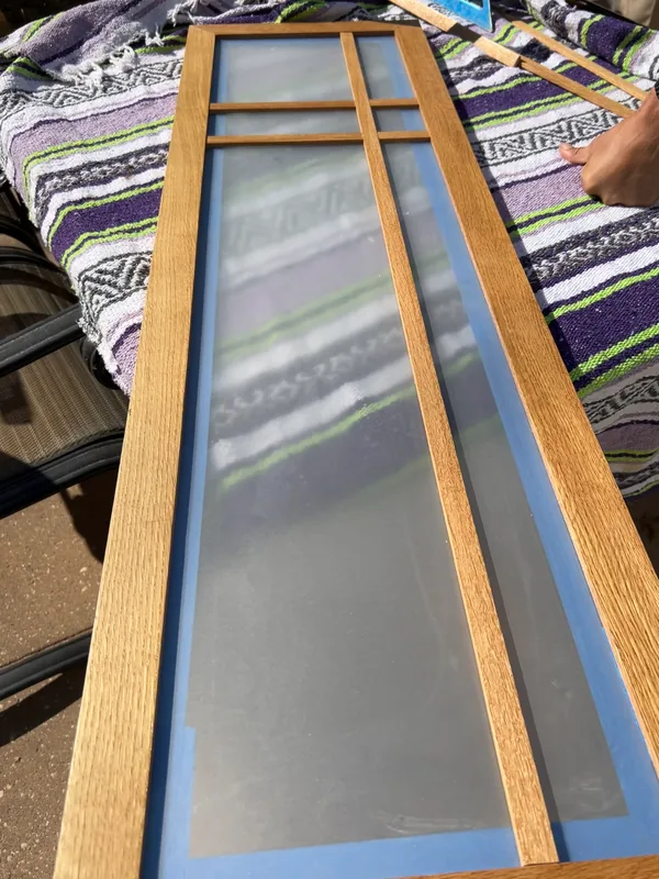 The custom shoji screen door lying on a work blanket during fabrication, showing the mitered oak frame and asymmetric grid before the frosted panel is set