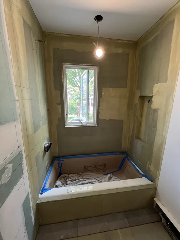 The new soaking tub set in the waterproofed alcove at rough-in, protected with board and tape, with the window above awaiting its shoji screen