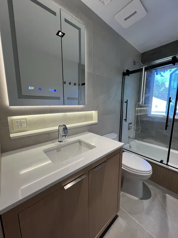 The finished Great Neck bathroom with custom vanity, backlit mirror, toilet and bypass glass tub enclosure