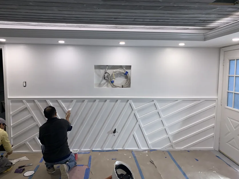 A carpenter kneeling on protected flooring, installing the custom white geometric millwork below the recessed media niche.