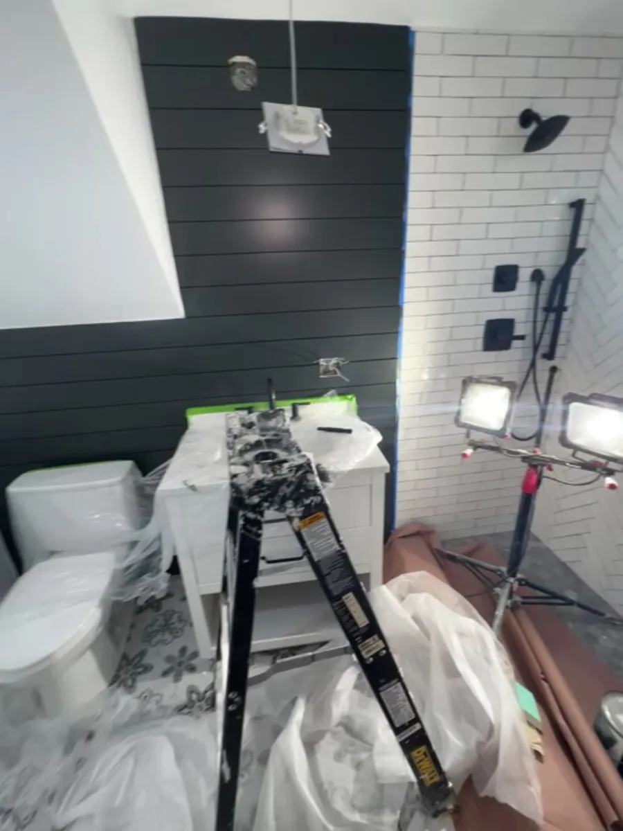 Mid-build: dark shiplap wall painted, white subway tile set in the shower, toilet and vanity roughed in, ladder in the room.
