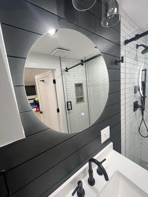 Round LED mirror on the dark shiplap wall with the clear-glass sconce above.