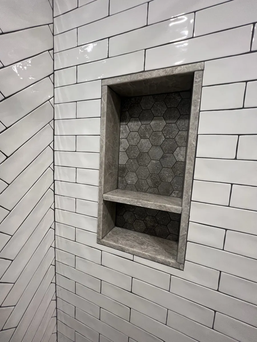 Close-up of the shower niche: marble-look stone surround and shelf, grey hex tile back, set into white subway and herringbone tile.