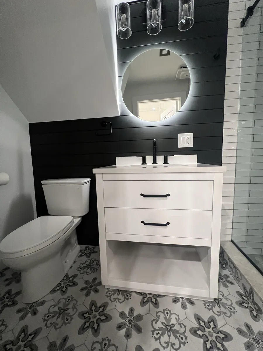 The finished room from the door: toilet and vanity under the sloped stair ceiling, dark shiplap wall, round LED mirror and the shower beyond.