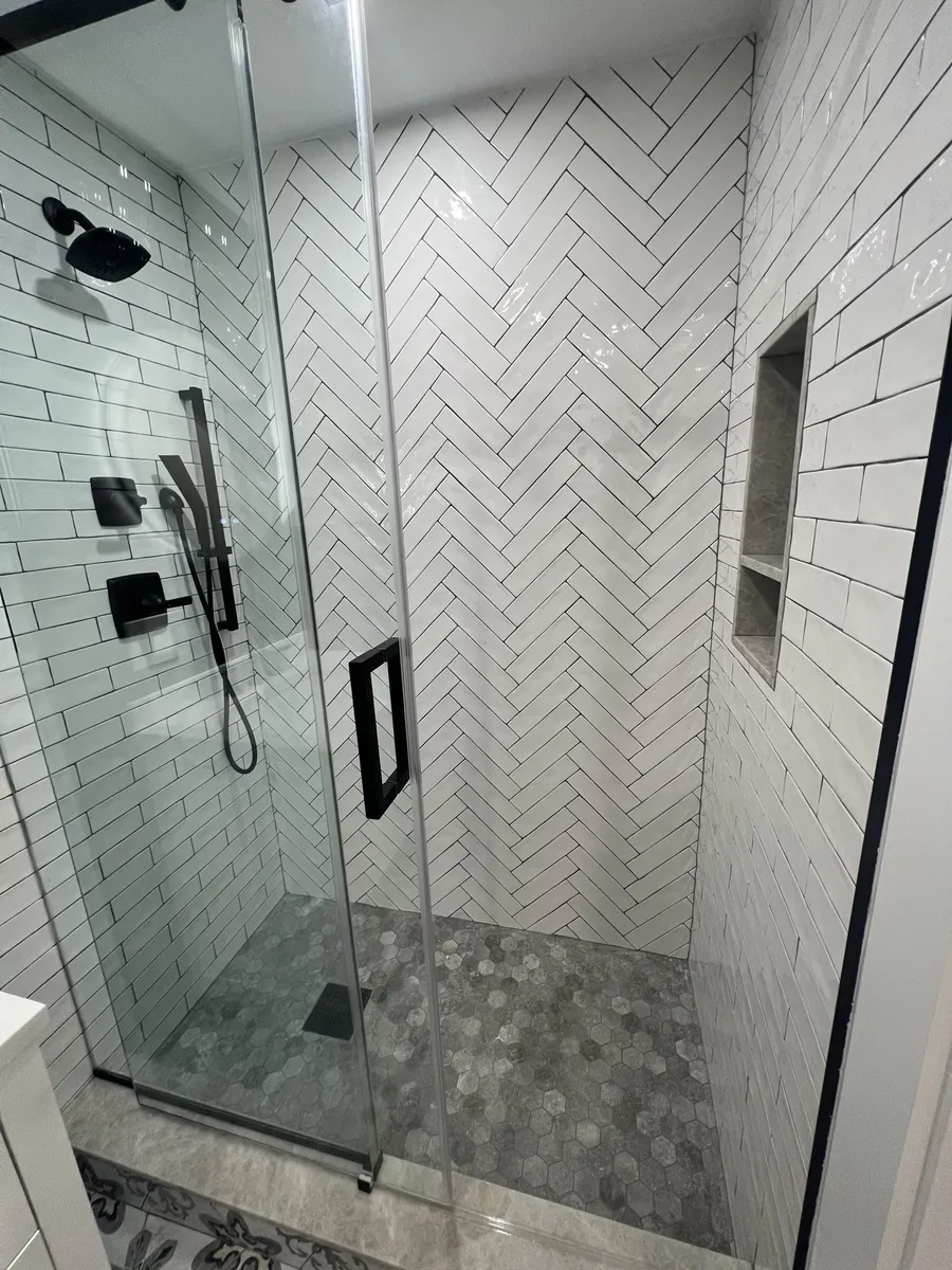 Inside the finished shower: herringbone subway feature wall, grey pebble hex floor, matte-black rain head, handheld slide bar and a tiled niche.