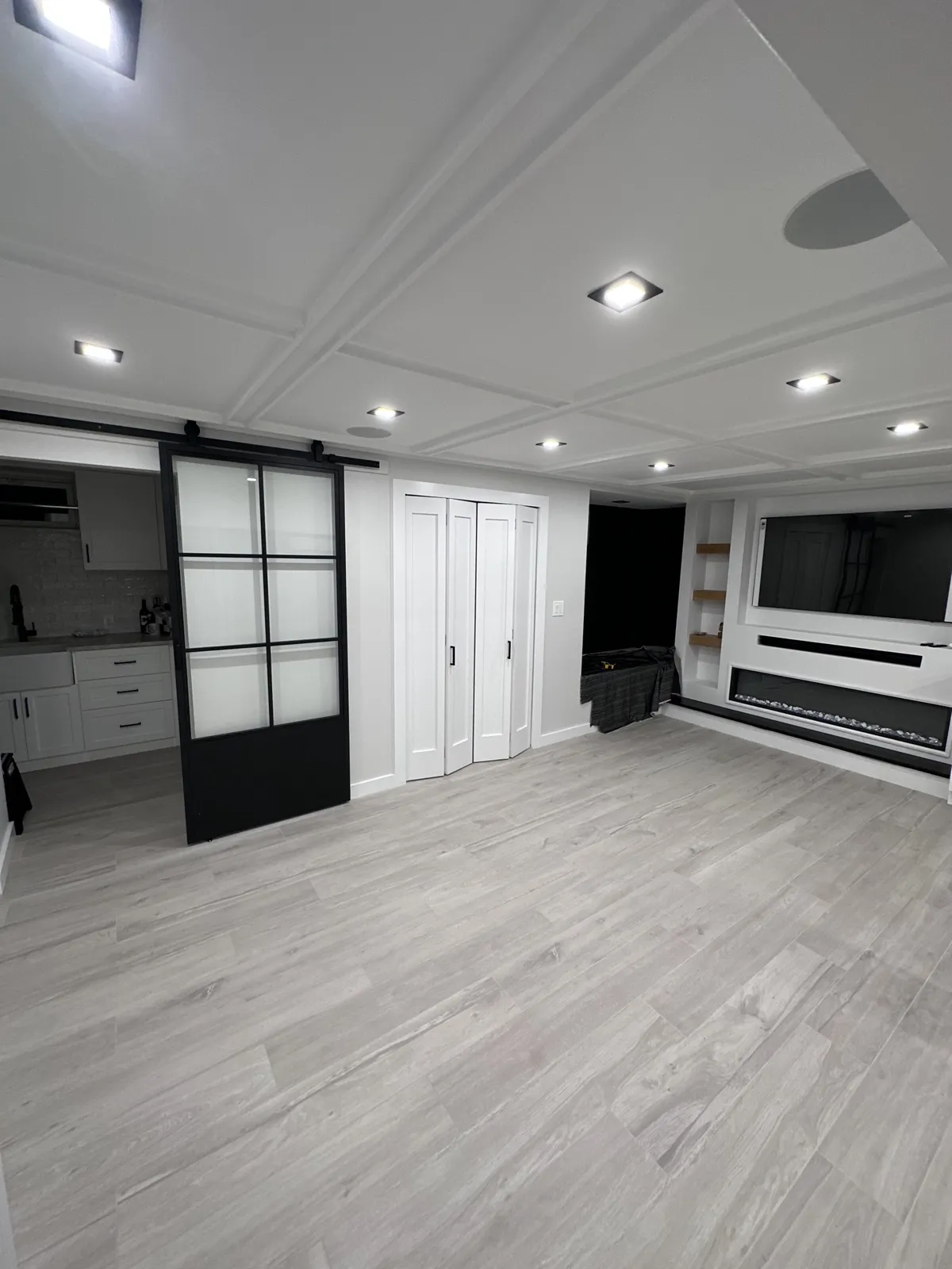 The finished basement from the far end: coffered ceiling with recessed lights, black-framed glass sliding door, double doors, and the media wall with linear fireplace on the right.