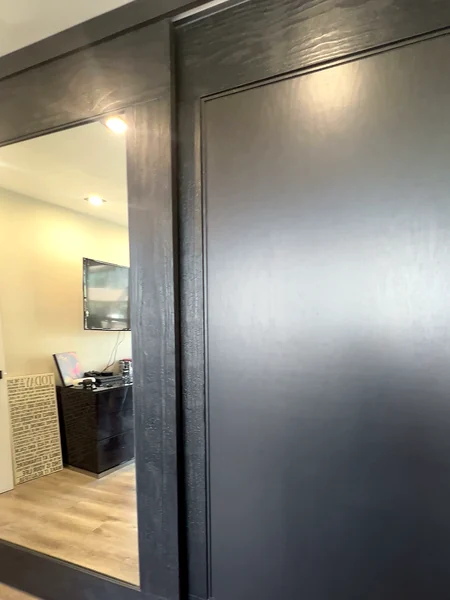 Oversized charred cedar closet door beside a full-height mirror panel that reflects the media wall across the room, capped by a charred cedar header, in Malba, Queens