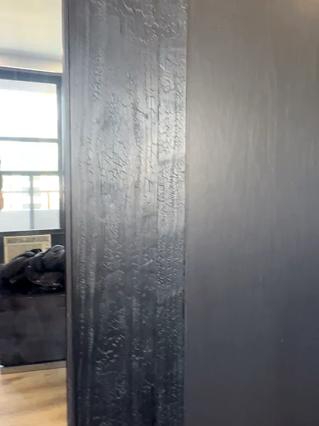 Window light raking across the cracked charred surface of the cedar closet door stiles beside the full-height mirror